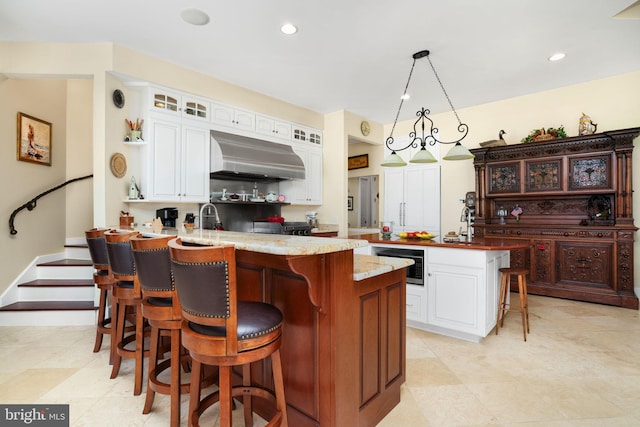 kitchen with decorative light fixtures, a peninsula, wall chimney range hood, light stone countertops, and glass insert cabinets