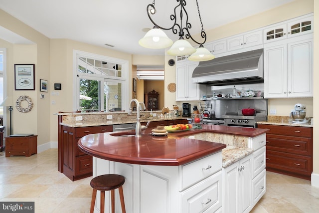 kitchen featuring glass insert cabinets, decorative light fixtures, a center island with sink, and wall chimney range hood