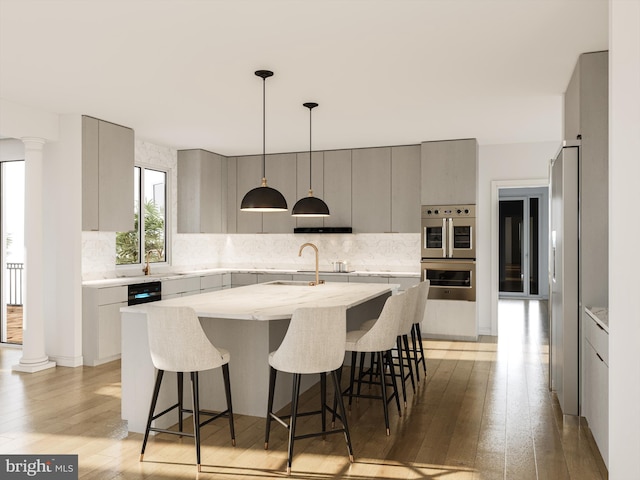 kitchen featuring backsplash, decorative light fixtures, stainless steel double oven, light hardwood / wood-style flooring, and a center island with sink