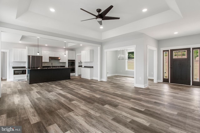 unfurnished living room featuring dark wood-type flooring, ceiling fan with notable chandelier, and a tray ceiling