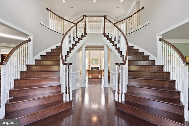 stairway featuring wood finished floors, decorative columns, a lit fireplace, a towering ceiling, and crown molding