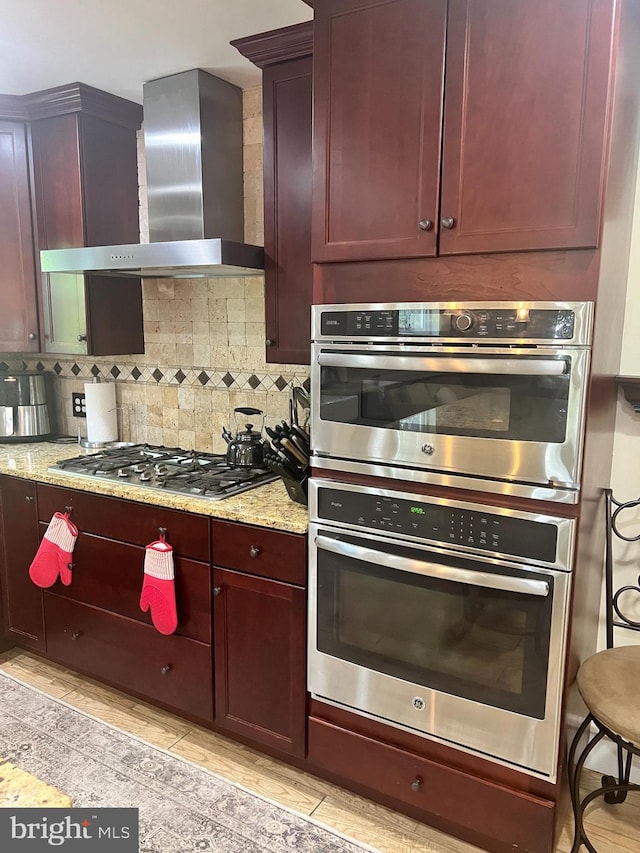 kitchen featuring stainless steel appliances, light stone counters, light hardwood / wood-style flooring, decorative backsplash, and wall chimney range hood