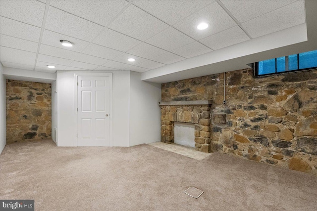 basement with a drop ceiling, a stone fireplace, and carpet floors