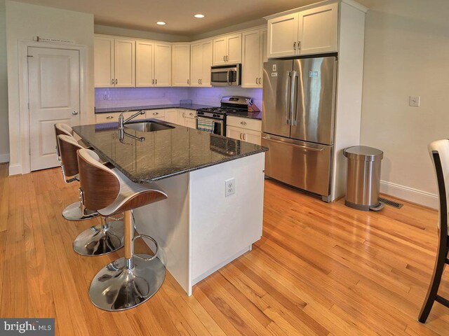 kitchen featuring appliances with stainless steel finishes, a kitchen island with sink, light wood-type flooring, and dark stone counters