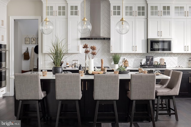 kitchen with appliances with stainless steel finishes, white cabinetry, a kitchen breakfast bar, hanging light fixtures, and wall chimney exhaust hood