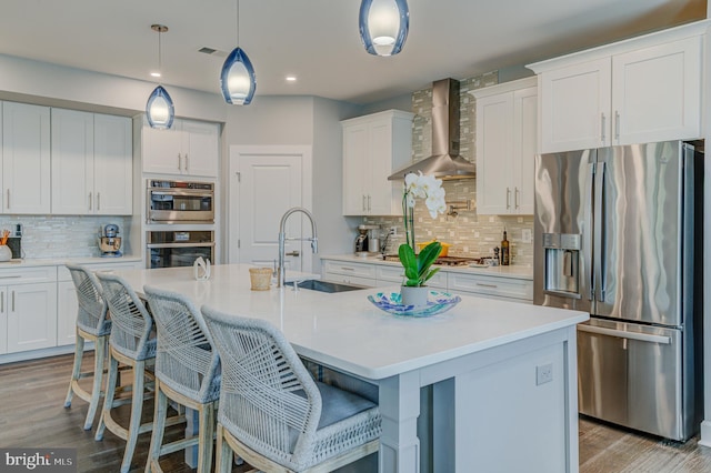 kitchen featuring white cabinets, appliances with stainless steel finishes, an island with sink, and wall chimney range hood