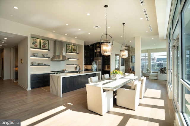 kitchen with sink, a center island with sink, light wood-type flooring, wall chimney range hood, and hanging light fixtures