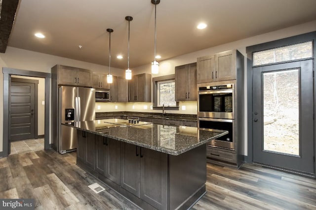 kitchen with stainless steel appliances, dark wood-style flooring, a center island, dark stone counters, and decorative light fixtures