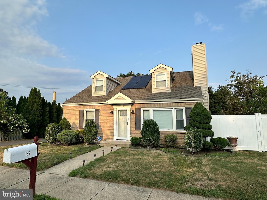 cape cod house featuring a shingled roof, a chimney, fence, roof mounted solar panels, and a front yard