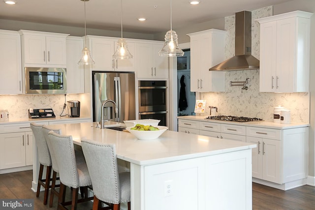 kitchen featuring white cabinetry, dark wood-type flooring, wall chimney exhaust hood, appliances with stainless steel finishes, and a center island with sink