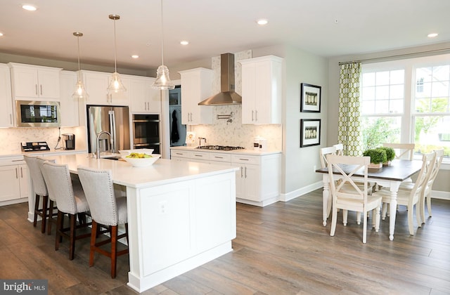 kitchen featuring a center island with sink, appliances with stainless steel finishes, dark wood-type flooring, white cabinetry, and wall chimney range hood