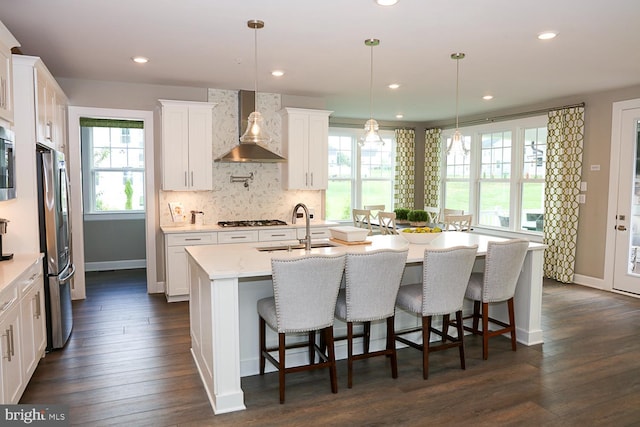 kitchen with wall chimney exhaust hood, dark hardwood / wood-style flooring, appliances with stainless steel finishes, an island with sink, and white cabinetry