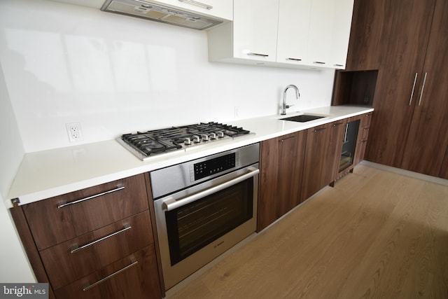 kitchen with sink, appliances with stainless steel finishes, white cabinetry, range hood, and light wood-type flooring