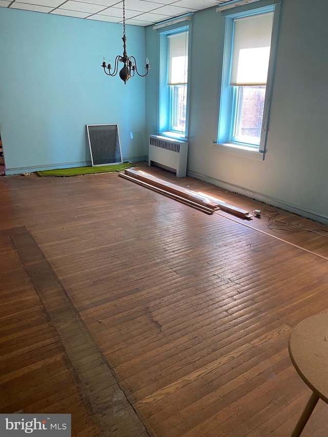 unfurnished dining area featuring radiator, hardwood / wood-style floors, a drop ceiling, and a chandelier