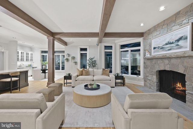 living room featuring beam ceiling, sink, decorative columns, light wood-type flooring, and a stone fireplace