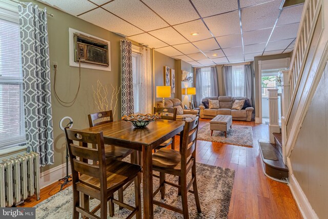 dining space featuring radiator, a wall mounted air conditioner, hardwood / wood-style floors, and a drop ceiling