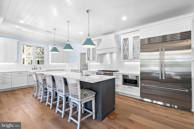 kitchen featuring an island with sink, custom range hood, glass insert cabinets, stainless steel appliances, and white cabinetry