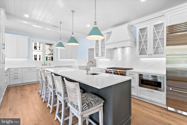 kitchen with an island with sink, glass insert cabinets, appliances with stainless steel finishes, custom exhaust hood, and a sink