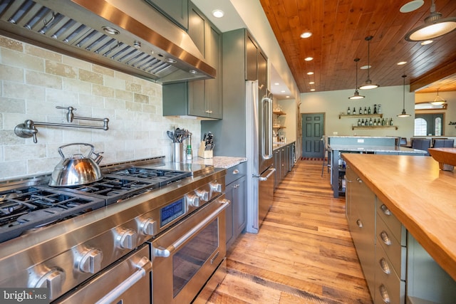 kitchen with backsplash, high end appliances, wall chimney range hood, pendant lighting, and light hardwood / wood-style floors