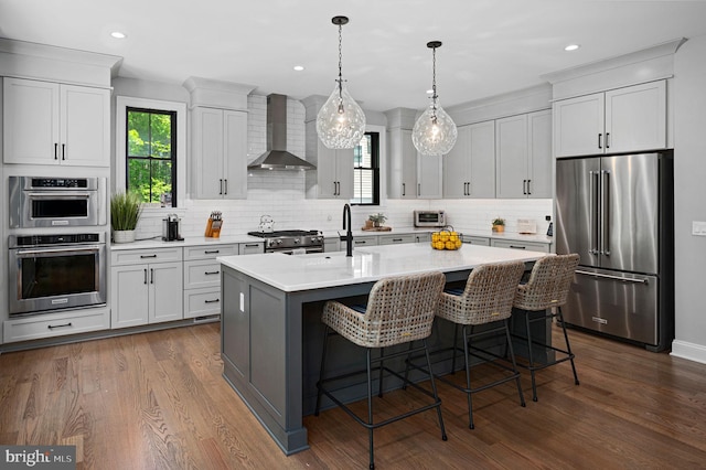 kitchen with dark wood-type flooring, a kitchen island with sink, high end appliances, decorative light fixtures, and wall chimney exhaust hood