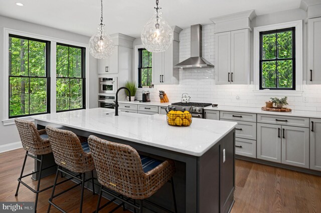 kitchen featuring appliances with stainless steel finishes, an island with sink, a kitchen breakfast bar, light stone countertops, and wall chimney exhaust hood