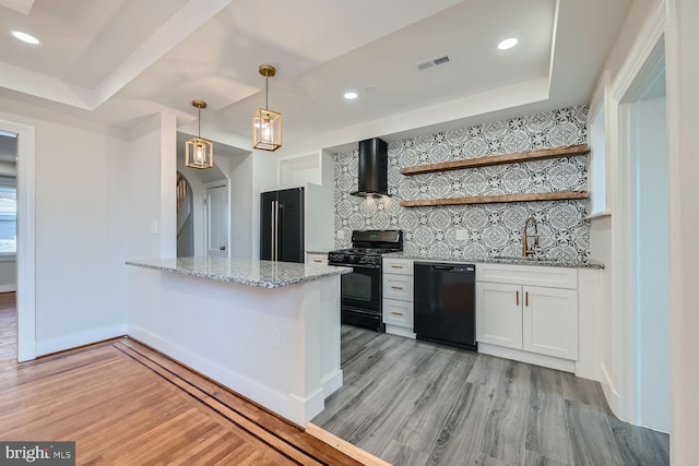 kitchen with light hardwood / wood-style floors, white cabinets, kitchen peninsula, black appliances, and wall chimney range hood