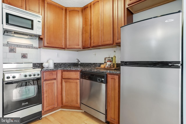 kitchen with appliances with stainless steel finishes, sink, dark stone countertops, backsplash, and light hardwood / wood-style flooring