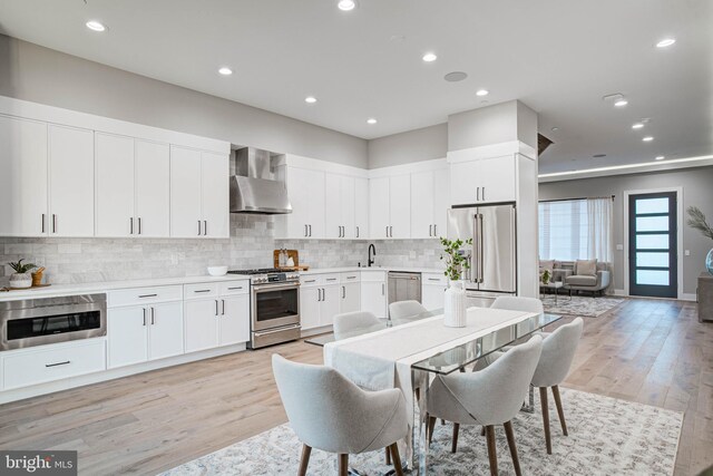 kitchen with light hardwood / wood-style flooring, decorative backsplash, stainless steel appliances, wall chimney range hood, and white cabinets