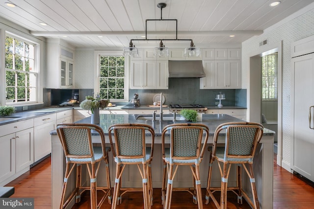 kitchen with wood-type flooring, a center island with sink, range hood, and hanging light fixtures