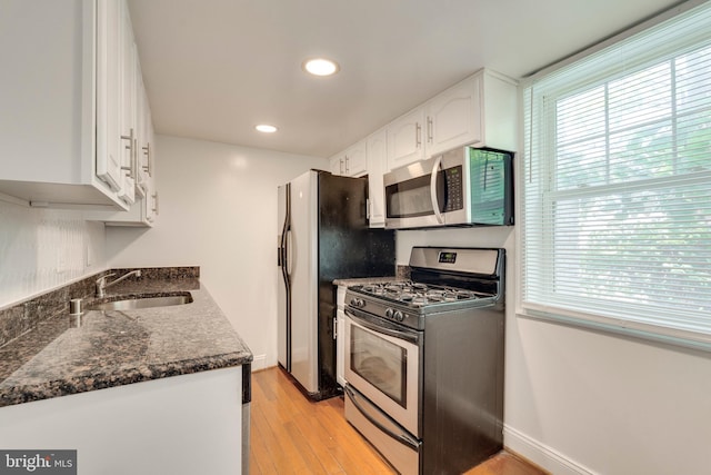 kitchen with light wood finished floors, stainless steel appliances, white cabinetry, a sink, and dark stone countertops