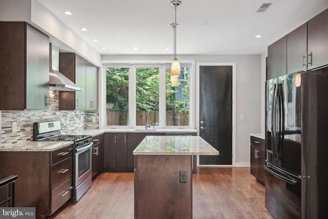 kitchen featuring wall chimney range hood, stainless steel gas range, black refrigerator, a kitchen island, and decorative light fixtures