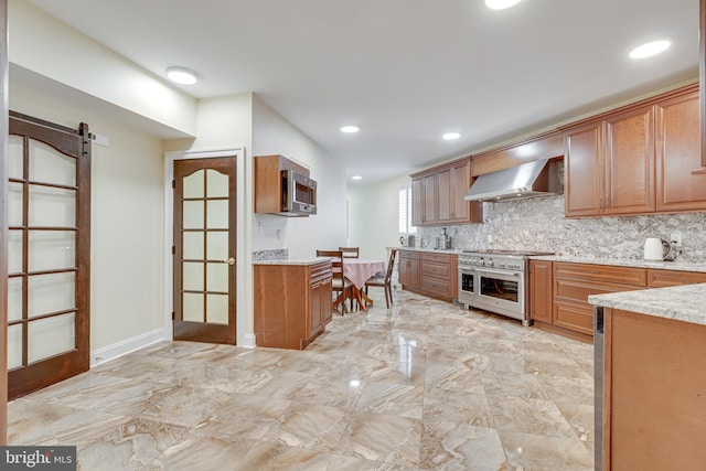 kitchen with wall chimney range hood, tasteful backsplash, a barn door, light stone counters, and stainless steel appliances