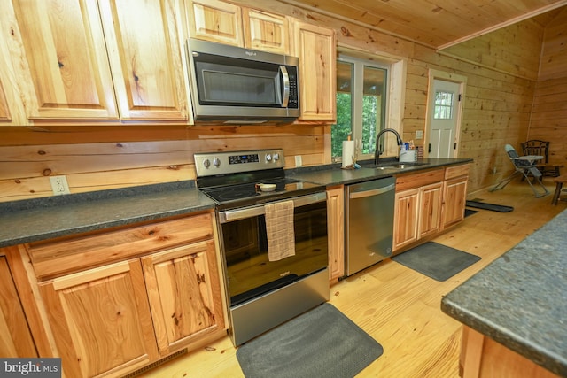 kitchen with light wood-style flooring, wooden walls, stainless steel appliances, a sink, and dark countertops