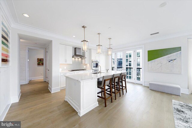 kitchen with backsplash, light hardwood / wood-style flooring, wall chimney exhaust hood, a kitchen island with sink, and white cabinets