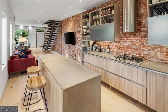 kitchen featuring stainless steel gas cooktop, light hardwood / wood-style flooring, brick wall, light brown cabinets, and a kitchen breakfast bar