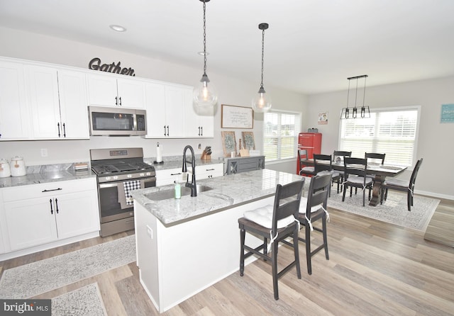 kitchen featuring a kitchen island with sink, sink, appliances with stainless steel finishes, decorative light fixtures, and white cabinetry