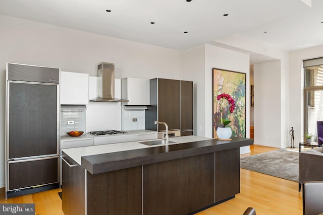 kitchen with light wood-type flooring, sink, dark brown cabinetry, paneled fridge, and wall chimney exhaust hood