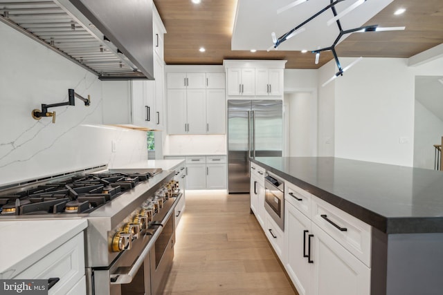 kitchen featuring decorative backsplash, built in appliances, white cabinets, and wall chimney exhaust hood