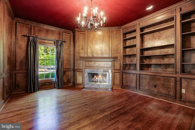 unfurnished living room featuring wood walls, a fireplace, a notable chandelier, wood-type flooring, and built in features