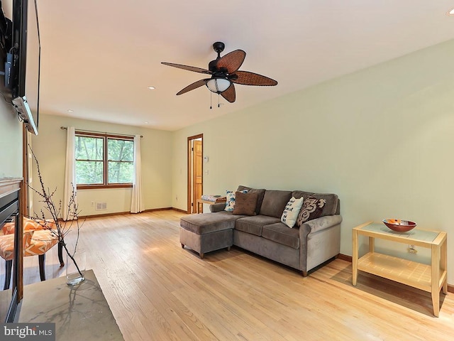 living area with light wood-type flooring, a glass covered fireplace, ceiling fan, and baseboards