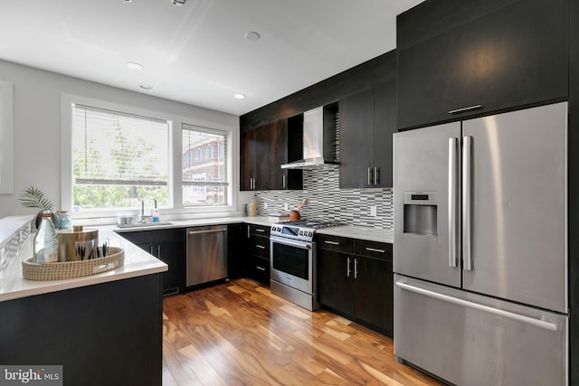 kitchen featuring wall chimney range hood, stainless steel appliances, sink, light wood-type flooring, and tasteful backsplash