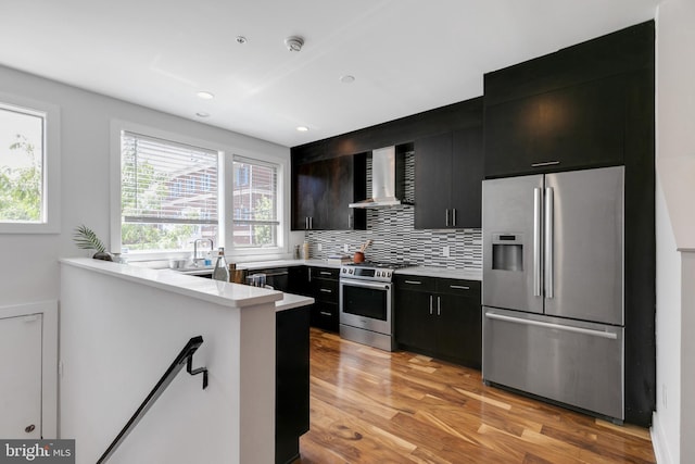 kitchen featuring decorative backsplash, wall chimney exhaust hood, kitchen peninsula, stainless steel appliances, and light hardwood / wood-style floors