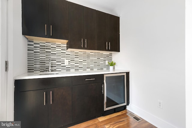 bar featuring decorative backsplash, wine cooler, sink, dark brown cabinetry, and light wood-type flooring