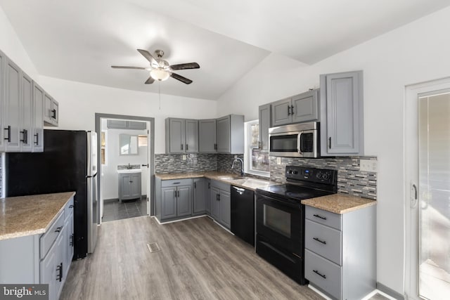 kitchen with vaulted ceiling, black appliances, sink, gray cabinetry, and light hardwood / wood-style flooring