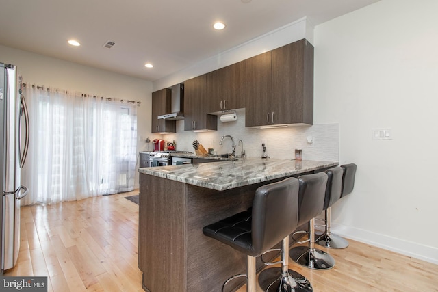 kitchen with wall chimney exhaust hood, light hardwood / wood-style flooring, stainless steel appliances, and light stone countertops