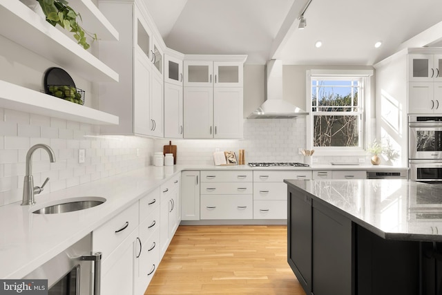 kitchen with open shelves, double oven, white cabinetry, a sink, and wall chimney range hood