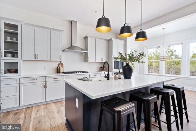 kitchen with a kitchen bar, decorative light fixtures, light hardwood / wood-style flooring, an island with sink, and wall chimney range hood