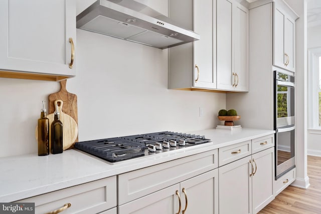 kitchen with light wood-type flooring, stainless steel double oven, wall chimney exhaust hood, gas cooktop, and white cabinets