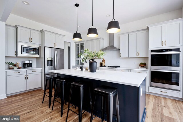 kitchen featuring wall chimney range hood, light wood-type flooring, a kitchen island with sink, a breakfast bar, and appliances with stainless steel finishes