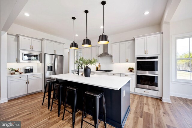 kitchen featuring light hardwood / wood-style floors, stainless steel appliances, wall chimney exhaust hood, and a center island with sink